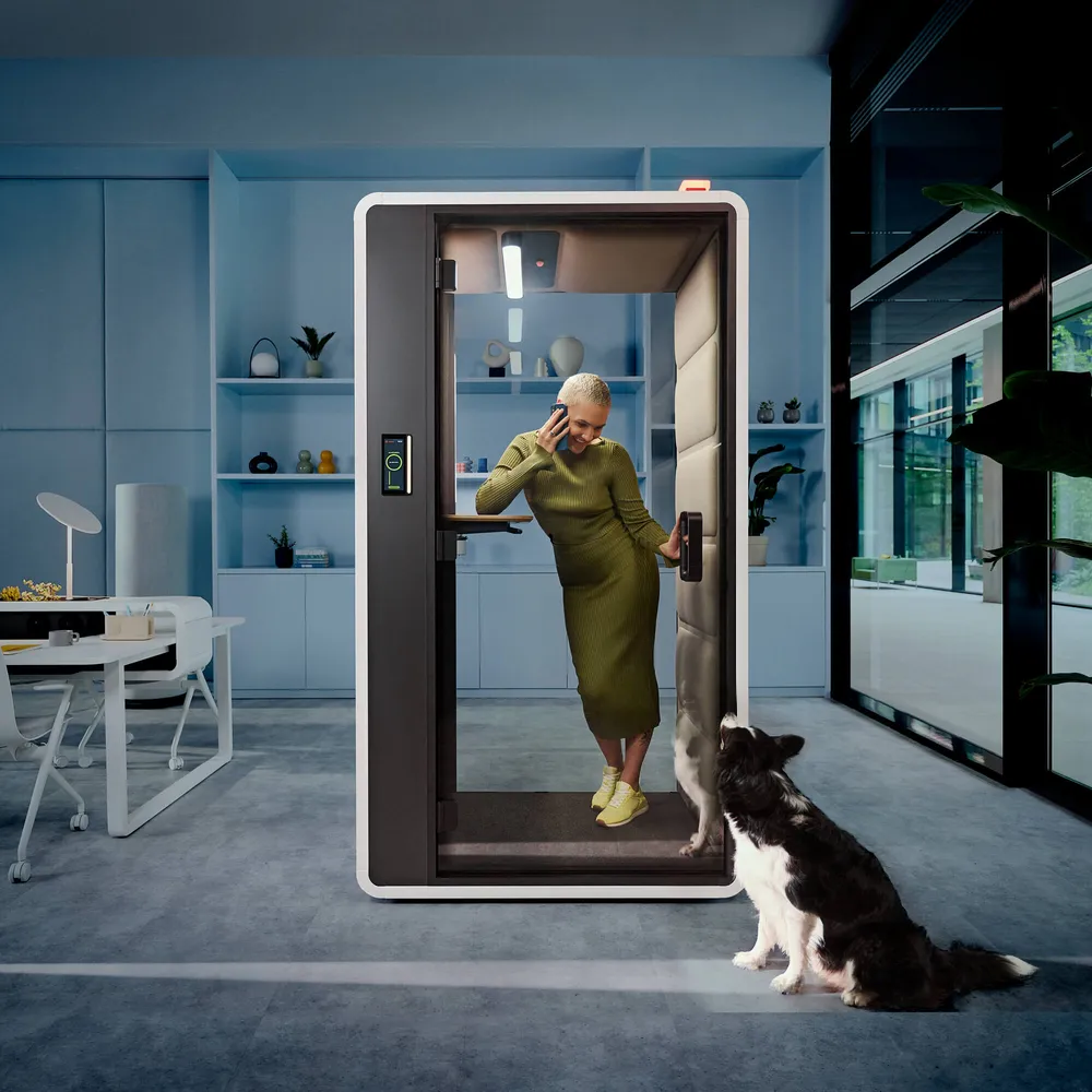 Een vrouw die staand aan het bellen is in een HushFreeS belcabine van Mikomax. Voor de belcabine zit een hond die contact maakt met de vrouw