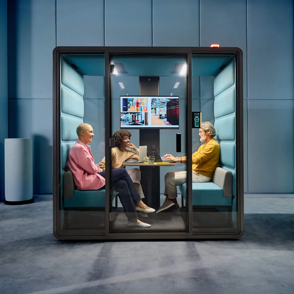 Een man die met twee vrouwen aan het vergaderen is in een HushFreeM vergadercabine van Mikomax welke in een donkere omgeving staat.